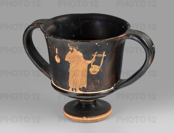 Red-figure kantharos with a depiction of an ephebe with an aryballos and a Greek inscription “Beautiful boy”., Kantharos with a slightly flared rim, deep bowl, flat bottom, high profiled stem, ring-shaped foot; thin-walled. A: bearded man with a lyre in left profile, holding a lyre in his left hand. B: ephebe with aryballos. Inner surface of the bowl covered with varnish. Technology: smooth surface; varnish thick, shiny, unevenly fired. Condition: vessel intact with small secondary losses and surface chips. Varnish color: 10 YR 2/1 black. Circa 450 BC mid-5th century BC. Attica. Type: kantharos vessel. Dimensions: height 11 cm; width 16.2 cm; depth 10.2 cm., A black pottery cup with a wide bowl, two curved handles, and a pedestal base, showing a reddish-orange painted figure of a robed person holding a stringed instrument and standing beside another hanging object; the cup has worn spots and small chips revealing lighter clay beneath, with visible colors including black, reddish-orange...