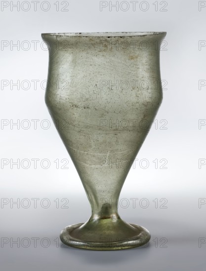 Chalice with a strongly elongated bowl widening upwards to 2/3 of its height, slightly recessed above below the flared rim. Wide foot with a ring-shaped rim. Translucent light green glass with visible streaks in the mass, uneven rim, cold-worked; foot with a clearance, made by pressing the bottom, free-blown. Type: Isings form 109b, Morin-Jean form 111. 4th century, Gaul France. Glass vessel chalice. Weight 0.158 kg; diameter overall 7.6 cm; foot diameter 4.5 cm., A single translucent glass vessel with a flared rim, a rounded shoulder narrowing into a conical body that tapers to a short stem and a circular foot, showing surface irregularities and tiny bubbles; the glass appears pale green with areas of darker green and subtle brownish spots, set against a neutral light gray background.