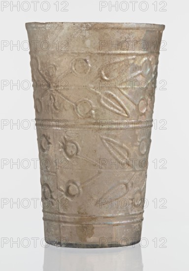 Glass decorated with a leaf and fruit motif; transparent, colorless glass with relief decoration in two registers separated by two thin horizontal convex lines; upper register bounded above by a single convex line, lower register bounded below by two horizontal convex lines; blown in a mold. Bottom diameter 4.25 cm, weight 0.066 kg. Dated 1st–2nd century previously dated 2nd century BC. Syria manufacture, Gaul finding., Vessel., A single cylindrical vessel photographed against a plain light background, with a slightly flared rim and a straight body that tapers subtly toward the base; the surface shows a raised repeating pattern of circular and elongated teardrop motifs arranged in horizontal bands separated by narrow ridges, and the texture appears rough and weathered with areas of iridescent sheen. The visible colors are muted beige and tan overall with patches of pale brown and subtle greenish and silvery iridescence.