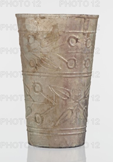 Glass decorated with a leaf and fruit motif;, transparent, colorless glass with relief decoration in two registers separated by two thin horizontal convex lines; upper register bounded above by a single convex line, lower register bounded below by two horizontal convex lines; blown in a mold. Bottom diameter: 4.25 cm; weight: 0.066 kg; dated 1st–2nd century previously dated 2nd century BC; manufactured in Syria; found in Gaul France., A single translucent cup with a tapered cylindrical shape stands upright against a plain pale background, showing an iridescent surface with a mix of muted beige, pale pink, light brown, and subtle greenish and silvery highlights; the exterior has decorative raised patterns forming repeating floral or leaf-like motifs and circular impressions arranged in horizontal bands, with a few parallel ridges encircling the top and bottom areas; the surface appears weathered with uneven texture, patches of cloudiness, and slight surface pitting, and the cup casts a ...