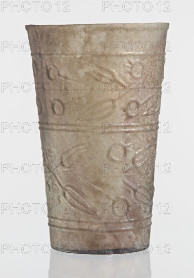 Glass decorated with a leaf and fruit motif;, Goluchów Greater Poland Voivodeship; transparent, colorless glass; relief decoration in two registers separated by two thin horizontal convex lines, upper register bounded above by a single convex line, lower register bounded below by two convex horizontal lines; blown in a mold. Bottom diameter: 4.25 cm; dated 1st–2nd century previously dated 2nd century BC; Syria manufacture; found in Gaul France; vessel; weight 0.066 kg., A single cylindrical cup with a slightly flared rim, shown upright against a pale background, made of translucent material with a weathered, iridescent surface in shades of beige, taupe, and muted green with subtle pinkish and gray undertones; the exterior bears repeated raised decorative motifs of circular and elongated almond-shaped elements arranged in horizontal bands separated by raised lines, and the base shows a darker rim and a faint reflective patch on the surface below.