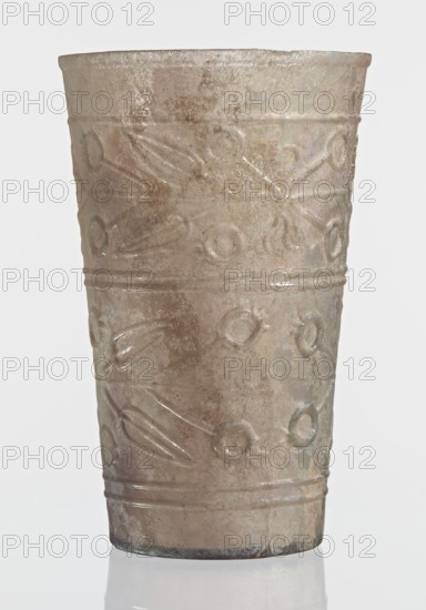 Glass decorated with a leaf and fruit motif;, Goluchów Greater Poland Voivodeship; transparent, colorless glass; relief decoration in two registers separated by two horizontal thin convex lines, the upper register bounded from the top by a single convex line, the lower register bounded from the bottom by two convex horizontal lines; blown in a mold. Bottom diameter: 4.25 cm; dated 1st–2nd century previously dated 2nd century BC; Syria manufacture, Gaul finding, France; vessel; weight 0.066 kg., A single cylindrical cup with a slightly flared rim standing upright on a reflective surface; the cup has a textured, weathered appearance with an iridescent sheen and a palette of muted beige, tan, brown, and subtle greenish and silvery highlights; the surface is decorated with a repeating embossed pattern of circular motifs connected by radiating petal-like shapes and horizontal banding near the top and bottom; the rim shows a rough, irregular edge and the base appears darker with some accumul...