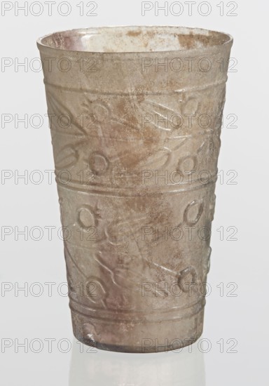 Glass decorated with a leaf and fruit motif, unknown creator; Goluchów Greater Poland Voivodeship; glass vessel with plant motifs; transparent, colorless glass; relief decoration in two registers separated by two horizontal thin convex lines; upper register bounded from the top by a single convex line; lower register bounded from the bottom by two convex horizontal lines; blown in a mold. Bottom diameter: 4.25 cm; dated 1st–2nd century previously dated 2nd century BC; manufactured in Syria; found in Gaul France; vessel; weight 0.066 kg., A tall cylindrical cup with a slightly flared rim, made of translucent glass showing a weathered, iridescent surface with shades of pale brown, gray, and subtle green and pink tones; the exterior features a repeating raised pattern of circular and linear motifs arranged in horizontal bands, with a worn and pitted texture and areas of cloudiness and spotting, and the cup sits on a reflective surface against a light neutral background.