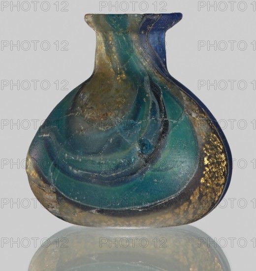 Unguentarium with carnation and a gold ribbon; bulging belly. 1st half of the 1st century BC. Type: Isings form 7. Mosaic glass small bottle with a cylindrical neck; sagging. Outer diameter of the spout: ? cm. Height: 2.9 cm. Body diameter: 2.6 cm. Bottom diameter: ? cm. Unknown workshop/creator, unknown place of origin. Vessel. Weight: 0.008 kg., A small, rounded glass vessel with a short neck and flared rim sits against a pale gray background, showing swirling bands of deep teal, turquoise, and dark blue with areas of greenish-blue; there are speckled patches of metallic gold and warm amber tones scattered across the surface, along with cloudy, translucent zones and thin pale veins where the glass appears worn or cracked, and the vessel casts a soft reflection beneath it.