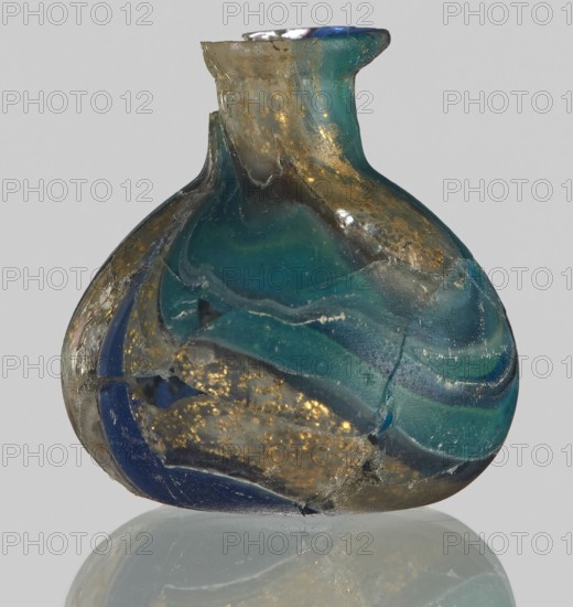 Unguentarium with carnation with a gold ribbon; with a bulging belly. 1st half of the 1st century BC. Type: Isings form 7. Mosaic glass small bottle with a cylindrical neck; sagging. Outer diameter of the spout: ? cm. Height: 2.9 cm. Body diameter: 2.6 cm. Bottom diameter: ? cm. Unknown place of origin. Vessel. Weight 0.008 kg., A rounded glass vessel with a short neck and flared rim sits against a pale background, showing a mix of translucent teal, deep blue, and green glass bands with areas of cloudy clear glass and embedded metallic gold flecks; the surface appears cracked and weathered with visible chips and rough edges, and the object rests on a reflective base that creates a faint mirrored impression.