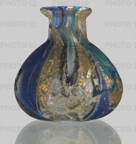Unguentarium with carnation with a gold ribbon; bulging belly. 1st half of the 1st century BC. Type: Isings form 7. Mosaic glass small bottle with a cylindrical neck; sagging. Height: 2.9 cm; Body diameter: 2.6 cm; Weight: 0.008 kg. Maker: Egyptian workshop, / unknown Italian workshop ., A small rounded glass vessel with a flared neck, showing a mix of deep blue, teal, gold, silver, and translucent pale areas, with irregular streaks and patches of color and some cloudy, textured inclusions and surface chips, sitting on a reflective surface that mirrors its colors.