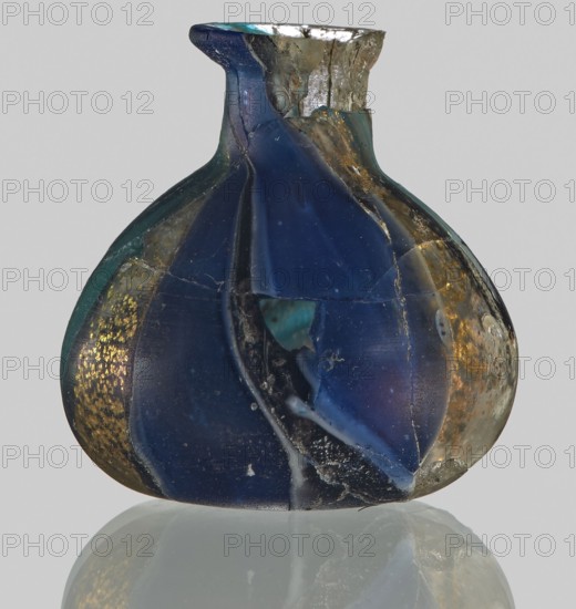 Unguentarium, Egyptian workshop ; unknown Italian workshop, attributed author unknown. Unguentarium with carnation with a gold ribbon; bulging belly. 1st half of the 1st century BC. Type: Isings form 7. Mosaic glass small bottle with a cylindrical neck; sagging. Outer diameter of the spout: ? cm. Height: 2.9 cm. Body diameter: 2.6 cm. Bottom diameter: ? cm. Vessel, height 2.9 cm, width 2.6 cm, weight 0.008 kg., A rounded glass vessel with a short neck and flared mouth sits against a light gray background, showing a few irregular chips and fractures. The body displays broad vertical segments of deep blue glass alternating with areas that reveal greenish-blue and translucent pale tones, and there are several metallic gold flecks and bands along some sections. The surface has a mix of smooth glossy areas and rougher worn patches with visible cracks and internal bubbles, and a faint reflection of the object appears beneath it.