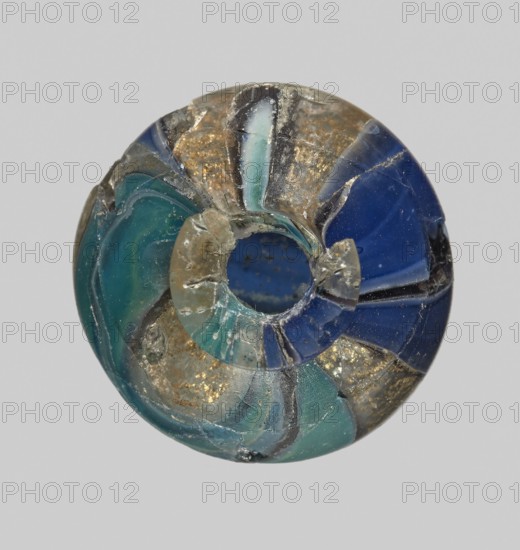 Unguentarium with carnation with a gold ribbon; with a bulging belly. Unguentarium. Mosaic glass small bottle with a cylindrical neck; sagging. Isings form 7. 1st half of the 1st century BC. Height: 2.9 cm; body diameter: 2.6 cm; weight: 0.008 kg. Maker: Egyptian workshop ; unknown Italian workshop, workshop attribution uncertain., A round, hollow object with a central hole showing a rough interior edge and a broken section, composed of curved segments in shades of blue and green with translucent and opaque areas, and shimmering flecks of gold-like material embedded in several segments; surface shows chips, cracks, and irregular textures, with visible layers and small reflective spots against a neutral light gray background.