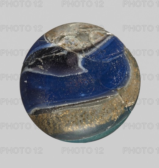 Unguentarium with carnation and a gold ribbon; bulging belly. Mosaic glass small bottle with cylindrical neck; sagging. Isings form 7. 1st half of the 1st century BC. Unguentarium. Height: 2.9 cm; body diameter: 2.6 cm; weight: 0.008 kg. Unknown workshop/creator; unknown place of origin., A round, glass-like object with a glossy surface showing a mix of deep blue, black, gray, green, gold-speckled, and beige areas; the surface has visible scratches, chips, and irregular edges, with smooth curved bands of color blending into one another and a few cloudy, translucent patches; small flecks and granular textures are visible within the gold-speckled region and along some of the worn edges.