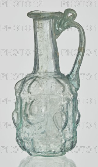Hexagonal jug, Goluchów Greater Poland Voivodeship. Hexagonal jug; eastern workshop; Syria country of find. 1st century AD. Type: Stern type with vessels, series A. Transparent, light green glass. Ovoid, hexagonal body decorated with a convex relief with a representation of vessels on the arms of an arcade with convex elements; cylindrical neck terminating in a flaring spout with an inwardly curved edge; blown in a three-part mold. Outer diameter of the spout: 1.90 cm. Height: 9.30 cm. Body diameter: 4 cm. Outer diameter of the bottom: 2.50 cm. 9.3 x 4 cm, 1st century, Syria, vessel, weight 0.034 kg., A translucent glass pitcher with a pale greenish-blue tint, featuring a narrow neck that widens into an irregular, bulbous body with raised, rounded protrusions and wavy contours; the handle is curved and attached near the rim with a decorative loop where it meets the neck, showing slight variations in thickness and small surface bubbles and imperfections throughout the glass; the rim is ...
