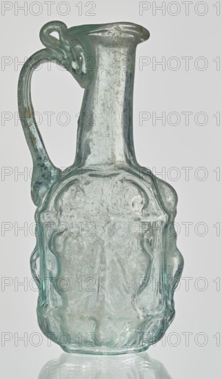 Hexagonal jug, Goluchów Greater Poland Voivodeship. Hexagonal jug; eastern workshop; Syria country of find; 1st century AD. Type: Stern type with vessels, series A. Transparent, light green glass. Ovoid, hexagonal body decorated with a convex relief with a representation of vessels on the arms of an arcade with convex elements; cylindrical neck terminating in a flaring spout with an inwardly curved edge; blown in a three-part mold. Outer diameter of the spout: 1.90 cm. Height: 9.30 cm. Body diameter: 4 cm. Outer diameter of the bottom: 2.50 cm. 9.3 x 4 cm. Vessel, weight 0.034 kg., A translucent glass pitcher with a pale green-blue tint, featuring a tall narrow neck that flares slightly at the rim and a curved handle attached near the top and midsection; the body is rounded with irregular, wavy raised ridges and molded decorative contours, showing areas of cloudiness and small surface imperfections, and the base reflects subtly on the surface below.