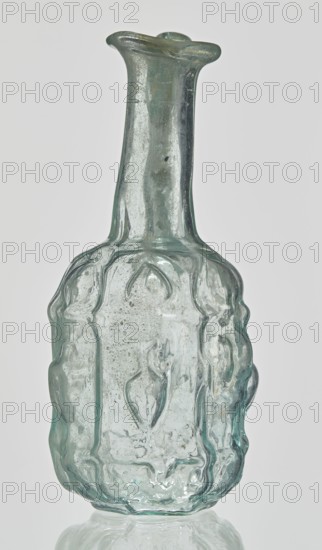 Hexagonal jug, Goluchów Greater Poland Voivodeship. Hexagonal jug; eastern workshop; Syria country of find; 1st century AD. Type: Stern type with vessels, series A. Transparent, light green glass. Ovoid, hexagonal body decorated with a convex relief with a representation of vessels on the arms of an arcade with convex elements; cylindrical neck terminating in a flaring spout with an inwardly curved edge; blown in a three-part mold. Outer diameter of the spout: 1.90 cm. Height: 9.30 cm. Body diameter: 4 cm. Outer diameter of the bottom: 2.50 cm. 9.3 x 4 cm, 1st century, Syria, vessel, weight 0.034 kg., A translucent glass bottle with a narrow neck and flared rim sits upright; the glass is a pale greenish-blue with areas of clear and cloudy texture, bubbles and irregularities visible throughout. The body of the bottle is bulbous with an embossed, sculpted surface featuring rounded vertical ridges and raised ornamental shapes that create a textured appearance. The neck is smoother and tap...