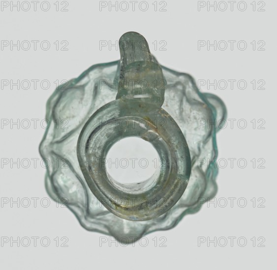 Hexagonal jug, eastern workshop, Syria country of find, 1st century AD. Stern type with vessels, series A. Transparent light green glass; ovoid hexagonal body decorated with a convex relief depicting vessels on the arms of an arcade with convex elements. Cylindrical neck terminating in a flaring spout with an inwardly curved edge; blown in a three-part mold. Outer diameter of the spout: 1.90 cm; height: 9.30 cm; body diameter: 4 cm; outer diameter of the bottom: 2.50 cm; weight: 0.034 kg., A translucent object with a pale green and aqua tint fills the frame, viewed from above showing a circular central opening surrounded by a rounded ring and an irregular scalloped outer edge; the glass-like material has cloudy areas, surface imperfections, and a few small brownish spots, and the overall shape includes a short looped protrusion at the top with similar greenish-blue coloration.