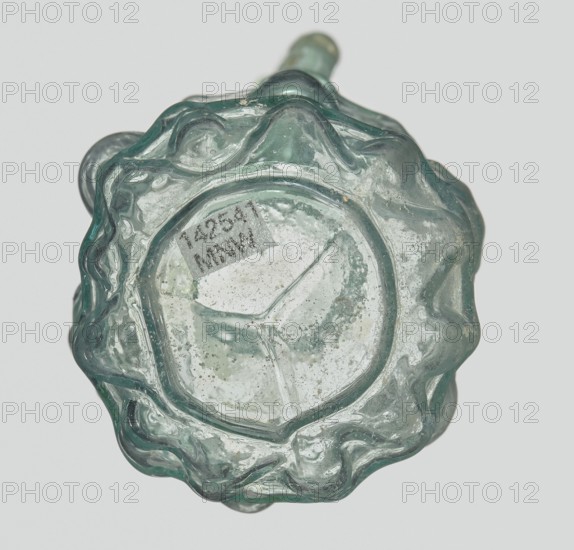 Hexagonal jug, Goluchów Greater Poland Voivodeship. Hexagonal jug; eastern workshop; Syria country of find, 1st century AD. Type: Stern type with vessels, series A. Transparent light green glass. Ovoid, hexagonal body decorated with a convex relief showing vessels on the arms of an arcade with convex elements. Cylindrical neck terminating in a flaring spout with an inwardly curved edge; blown in a three-part mold. Outer diameter of the spout: 1.90 cm. Height: 9.30 cm. Body diameter: 4 cm. Outer diameter of the bottom: 2.50 cm. 9.3 x 4 cm, weight 0.034 kg., A close-up top view of a glass object with an irregular scalloped rim, showing a pale green translucent color with areas of cloudy white deposits and small air bubbles; the surface has glossy highlights and some rough, textured spots, and the background is a very light gray.