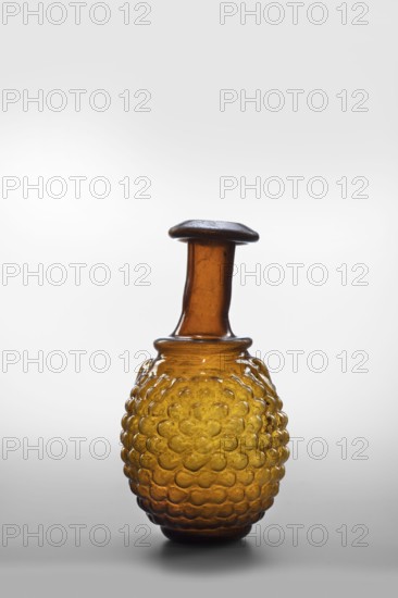 Bottle; with a bulging belly with decoration in the shape of a bunch of grapes; Syro-Palestine place of production. Translucent amber glass; body blown in a three-piece mold, neck and spout formed by free blowing. 3rd century; found in France; origin Syria. Vessel. Rim diameter 3.6 cm, base diameter 3.7 cm, weight 0.074 kg. Maker and workshop unknown., A single amber-colored glass bottle with a bulbous, textured body covered in a pattern of raised rounded bumps and a narrower neck topped by a flat, darkened rim, shown against a neutral light-gray background.