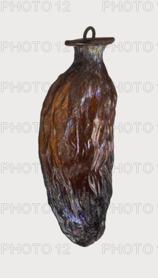 Date-shaped unguentarium; Syro-Palestine workshop; Smyrna place of discovery; 1st–beginning of 2nd century AD; Type: Isings form 78D. Translucent amber glass, inclined rim with inwardly curved edge; blown in a two-part mold. Rim diameter 2 cm; height 8.15 cm; belly diameter 2.95 cm; weight 0.020 kg., A single elongated object with an irregular, textured surface resembling a vessel or pod, predominantly brown with areas of dark brown and black; the surface shows iridescent highlights in purple, blue, and hints of green and pink; the top narrows into a short neck with a flat, circular rim and a small loop or handle attached, both darker brown to black; the body has flowing, vertical ridges and a glossy, slightly reflective finish interspersed with matte, rougher patches.