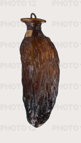 Date-shaped unguentarium; Syro-Palestine workshop; Smyrna place of discovery; 1st–beginning of 2nd century AD; Type: Isings form 78D. Translucent amber glass, inclined rim with inwardly curved edge; blown in a two-part mold; 1st century BC–beginning of 2nd century AD. Vessel, rim diameter 2 cm, height 8.15 cm, belly diameter 2.95 cm, weight 0.020 kg., The image shows a single elongated, irregularly shaped object resembling a bottle or vessel with a narrow neck and flared rim, suspended against a pale background. The surface has a rough, textured appearance with vertical ridges and an uneven finish. Visible colors include various shades of brown, amber, dark brown to near-black, and subtle hints of iridescent blues and purples in places. A small loop or handle sits atop the rim in a dark color. The object appears glossy in some areas and matte in others, with lighter amber tones concentrated near the neck and darker tones toward the lower portion.