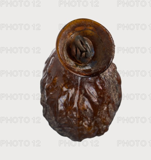 Date-shaped unguentarium; Syro-Palestine workshop; Smyrna place of discovery; 1st–beginning of 2nd century AD; Type: Isings form 78D. Translucent amber glass, inclined rim with inwardly curved edge; blown in a two-part mold; 1st century BC–beginning of 2nd century AD; vessel. Rim diameter 2 cm; height 8.15 cm; belly diameter 2.95 cm; weight 0.020 kg., A close-up of a single irregularly shaped object with a glossy, wrinkled surface in shades of brown and amber, showing darker brown and blackened areas and some lighter brown highlights; the top of the object has a rounded opening with a ring-like element inside that appears darker and metallic in color, and the rim shows textured edges and amber tones; the background is a plain off-white or very light gray.