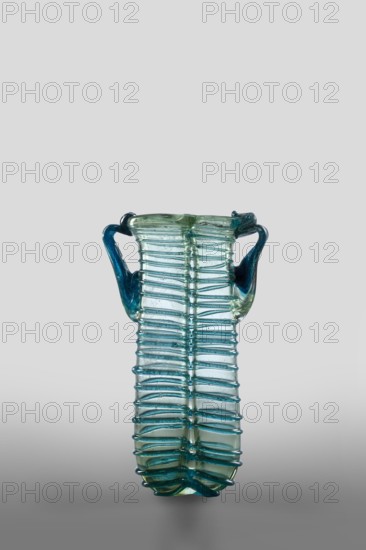 Dilekyt, unknown Syrian workshop; lekythos glass vessel, tube with spirally applied thread folded in half. Type: Barag type XII:2-1. Transparent, colorless glass with light bluish-green tint; decoration of spirally twisted translucent blue glass thread; free-blown. Outer rim diameters: 1 — 3.20 cm, 2 — 2.70 cm. 4th century. Vessel dimensions: height 11.7 cm, width 7.2 cm, depth 3.2 cm; weight 0.08 kg., A translucent glass vessel with a tall, elongated body and a pair of curved handles, showing a few shades of blue and green; the body has a ribbed, ladder-like texture formed by several horizontal glass ridges wrapping around a central vertical seam, and the handles attach near the top and curve outward before returning inward, with the top rim slightly flared and uneven; the background is a smooth gradient of light gray to darker gray.
