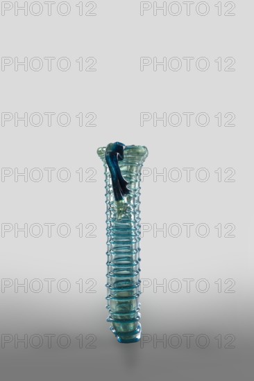 Dilekyt, unknown Syrian workshop; lekythos glass vessel, tube with spirally applied thread folded in half. Type: Barag type XII:2-1. Transparent, colorless glass with light bluish-green shade; decoration of spirally twisted translucent blue glass thread; free-blown. Outer diameter of the rim: 1 – 3.20 cm, 2 – 2.70 cm. 4th century, Vessel height 11.7 cm, width 7.2 cm, depth 3.2 cm, weight 0.08 kg., A tall translucent glass object stands upright against a neutral gray background, with a ribbed, cylindrical body featuring a repeating horizontal ring pattern and a flared top; the glass shows shades of aqua, teal, and blue with areas that appear lighter and more greenish, and a darker blue draped glass element rests over the top edge, extending partway down the side.
