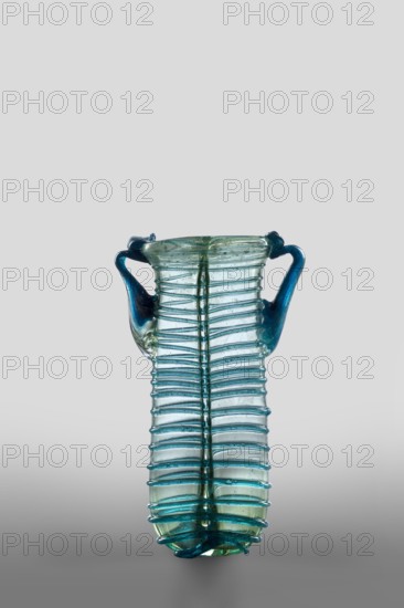 Dilekyt, unknown Syrian workshop; lekythos glass vessel, Barag type XII:2-1. Transparent, colorless glass with light bluish-green tint; decoration of spirally twisted translucent blue glass thread applied by free blowing; tube, spirally applied thread folded in half. Outer diameter of rim: 1 – 3.20 cm, 2 – 2.70 cm. 4th century; vessel height 11.7 cm, width 7.2 cm, depth 3.2 cm; weight 0.08 kg., A transparent glass vessel with a tall narrow body and a rounded base, featuring a ribbed texture of raised horizontal ridges running along its length and a vertical seam down the middle; the vessel has two curved handles attached near the rim that extend outward and up, and the glass shows shades of blue and green with areas of darker blue on the handles and rim and lighter greenish-blue tones on the body, set against a plain neutral gray background.