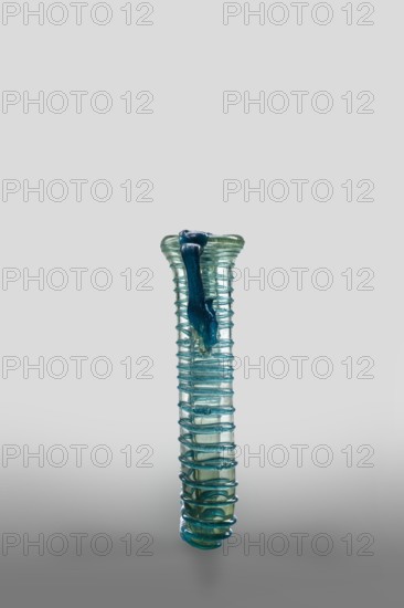 Dilekyt, unknown Syrian workshop,  workshop author; lekythos glass vessel, tube with spirally applied thread folded in half. Type: Barag type XII:2-1. Transparent, colorless glass in a light bluish-green shade; decoration of spirally twisted thread of translucent blue glass; free-blown. Outer diameter of rim: 1 – 3.20 cm, 2 – 2.70 cm. 4th century, unknown provenance. Vessel height 11.7 cm, width 7.2 cm, depth 3.2 cm, weight 0.08 kg., A tall, narrow glass vessel with a ribbed, spiral texture running along its length and a slightly flared top; the glass is translucent with shades of blue and green, and a darker blue blob of glass is attached near the rim, contrasting with the lighter aqua tones of the body; the background is a smooth gradient of light gray to darker gray at the bottom.