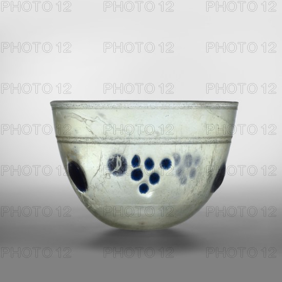 Bowl or lamp, hemispherical glass vessel, light green transparent glass, free-blown, decorated with translucent cobalt glass discs placed in one line halfway up the bowl in six places in two alternating arrangements 1 — a single large disc; 2 — six smaller ones arranged like a grape. Decorated with knobs. Isings form 106 96A. Outer rim diameter 14.30 cm; height 9.30 cm; weight 0.306 kg. 4th century, Syria., A small translucent bowl with a pale greenish tint and a slightly iridescent surface, showing areas of cloudiness and fine surface scratches; the bowl has a rounded shape with a gently flaring rim and a horizontal band circling its upper portion; there are a few deep blue circular spots clustered on one side and a couple of larger dark blue oval spots spaced around the bowl; the background is a smooth gradient from light gray at the top to darker gray at the bottom, and the bowl sits on a soft gray surface.
