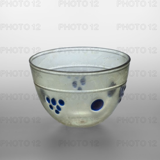 Bowl decorated with knobs, hemispherical glass bowl or lamp, decorated with cobalt oval discs; light green, transparent glass, decorative translucent cobalt discs placed in one line halfway up the bowl in 6 places, arranged alternately 1 — a single large disc, 2 — six smaller ones in a grape shape; free-blown. Type: Isings form 106 96A. Outer rim diameter: 14.30 cm; height: 9.30 cm; weight: 0.306 kg. 4th century, Syria., A translucent glass bowl with a rounded shape and a slightly flared rim, showing a pale greenish-gray color overall with areas of iridescent sheen, decorated with several deep blue circular spots and clusters of smaller blue dots around its exterior, and faint surface cracks and weathering marks visible on the glass.