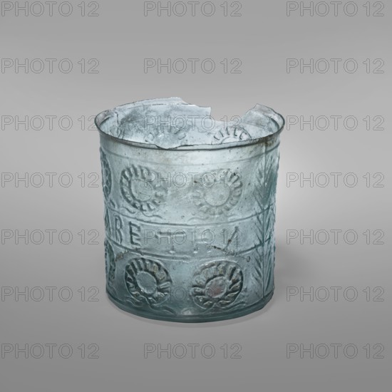 Glass with a wreath motif, Greek inscriptions. Transparent bluish-green glass decorative vessel, decorated in relief in three registers separated by horizontal rollers: upper and lower registers with wreath motifs; middle register with the inscription “LABE THN NEIKHN”; two opposite sides with vertical palm leaf motifs. Blown in a three-part form. Type: Harden group K.1.IIIk. Dating: mid-19th century classification; original date 1st century previously dated late 1st century BC–1st century AD. Likely origin: Sidon, Syria. Vessel; inscribed glass vessel, wreaths, palm trees, victories, leaves. Weight 0.048 kg; height 6.9 cm; diameter 7 cm; base diameter 6.3 cm., A translucent cylindrical glass object with an irregular broken rim, showing embossed circular floral patterns around its surface and banded horizontal ridges; the glass has a pale bluish-green tint with areas of cloudy white deposits and darker greenish spots, and the background is a smooth gradient of light gray.