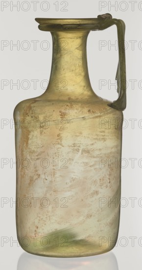 Jug, cylindrical body, high neck with flared spout and vertical vice; 2nd–3rd century AD; unknown eastern workshop; Goluchów Greater Poland Voivodeship; Syria; vessel; height 21.5 cm, width 10.5 cm., A translucent glass vessel with a narrow neck, flared rim, and a single curved handle attached near the top, showing a patina of cloudy and textured surface areas; colors visible include pale yellow, olive green, light beige, and hints of brown, with a glossy sheen and subtle streaks and smudges across its body.