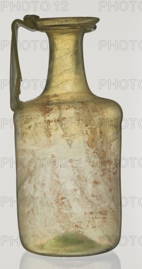 Jug, unknown eastern workshop, Cylindrical body, high neck with flared spout and vertical vice, 2nd–3rd century AD, Syria, vessel, height 21.5 cm, width 10.5 cm, A single glass vessel with a narrow neck flaring into a wide lip and a handle attached from the lip to the shoulder, shown upright against a plain light background; the glass appears mostly pale greenish-yellow with areas of iridescent sheen and patches of brownish discoloration across the body; the shape is squat and cylindrical toward the lower body with a gently curved shoulder and a more tapered neck, and the handle is slender and flat with a slight twisted form.