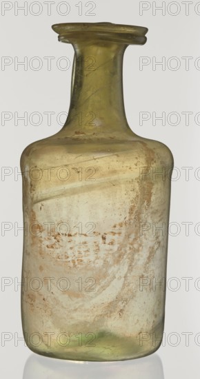 Jug, unknown eastern workshop, Cylindrical body, high neck with flared spout and vertical vice, 2nd–3rd century AD, Syria, vessel, height 21.5 cm, width 10.5 cm, The image shows a single translucent bottle with a narrow neck and a flared rim, made of glass that appears olive-green and pale yellow with areas of cloudy white and brownish surface deposits; the bottle has a rounded rectangular body with worn, uneven textures and subtle swirled patterns, and a few darker green and brown speckles and streaks visible across its surface, all set against a plain, light background.