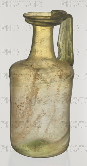 Jug. Cylindrical body, high neck with flared spout and vertical vice. 2nd–3rd century AD. Syria. Vessel. Height 21.5 cm; width 10.5 cm. Maker: unknown eastern workshop., A translucent glass vessel with a narrow neck and flared rim, showing a single handle attached from the rim to the shoulder; the glass is primarily pale yellow-green with areas of cloudy, muted green and brownish weathering; the surface shows irregular patches and streaks of earthy brown and tan discoloration, with a slightly iridescent sheen in places; the handle is a deeper green and appears thicker and slightly irregular in shape; overall the vessel has a worn, aged appearance against a plain light background.
