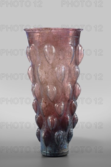 Glass, Mug glass vessel, Isings form 31. Transparent manganese iridescent glass, blown in a 4-part mold. Decoration: six rows of convex stepped elements shaped like almonds lotus buds; two concentric circles on the bottom. Outer diameter of bottom: 5 cm; weight 0.188 kg. Date: 2nd quarter of the 1st century. Findspot: Syria., A tall glass vessel with a tapered form, showing a translucent pink to purple hue that becomes darker toward the base, covered in a pattern of raised teardrop-shaped protrusions arranged in vertical rows; the surface shows areas of iridescent blue-green and darker brownish spots suggesting surface weathering, and the rim is slightly flared and uneven, all set against a plain light gray background.