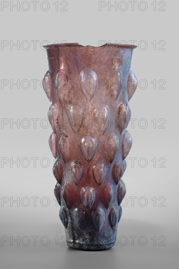 Glass, Mug glass vessel, Isings form 31. Transparent manganese iridescent glass, blown in a 4-part mold. Decoration: six rows of convex stepped almond-shaped elements lotus buds; two concentric circles on the bottom. Outer diameter of the bottom: 5 cm; weight 0.188 kg. Dated to the 2nd quarter of the 1st century, Syria., A tall vase with a textured surface covered in raised teardrop shapes, showing a mix of pink, purple, blue, and brown tones across its body, with areas of glossy and matte finish and some darker mottled spots; the vase tapers slightly toward the base and has an uneven rim with a small notch, and it is shown against a neutral light gray background.