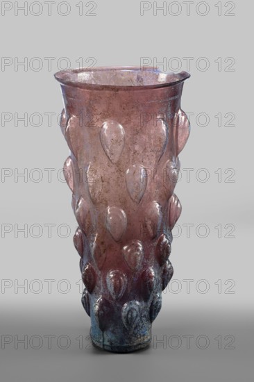 Glass mug; transparent, manganese, iridescent glass. Decorated with 6 rows of convex, stepped elements shaped like almonds lotus buds. Two concentric circles on the bottom; blown in a 4-part form. Isings form 31. Outer diameter of the bottom: 5 cm; weight 0.188 kg. Dated 2nd quarter of the 1st century, Syria., A single tall translucent vessel with a flared rim stands against a plain light gray background, showing a pinkish to purplish overall hue with areas of bluish-green iridescence and weathered discoloration; the surface is decorated with a repeated raised teardrop motif arranged in vertical rows, and the glass shows spots of mottling and patina with darker, more bluish tones concentrated near the base while the upper area remains more pink and slightly worn, and the rim has an irregular small chip.