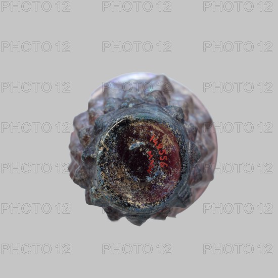 Glass, Mug glass vessel, Isings form 31. Transparent manganese iridescent glass, blown in a 4-part mold. Decoration: six rows of convex stepped almond-shaped elements lotus buds; two concentric circles on the bottom. Outer diameter of the bottom: 5 cm; weight 0.188 kg. Dated to the 2nd quarter of the 1st century, Syria., The image shows the top view of a textured object with a round central opening and a spiky, ridged outer surface. The object displays colors including dark blue, purple, pink, brown, and hints of green, with areas of lighter beige or off-white speckling. The background is a flat light gray.