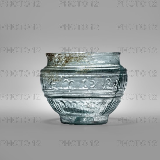 Glass bowl with Greek inscription. Transparent light-blue blown glass in a three-part form, cold-worked with an uneven rim edge. Relief decoration in the lower register: radially arranged long leaves flutes. In the upper, widest part of the bowl is a Greek inscription: Rejoice that you have come, bounded at top and bottom by horizontal ribs. Type: Harden group G.1.IIa. Dated 1st century Sidon; possibly associated with Cyprus. Vessel; weight 0.070 kg; diameter 4.5 cm bottom, diameter 7.4 cm rim; height 7 cm., A clear, translucent glass bowl with a pale green-blue tint sits centered on a neutral gray background, showing a rounded body with a slightly flared rim and a narrower base; the glass surface has a textured pattern of raised horizontal bands and decorative ridges around the middle and lower sections, and there are areas of darker brownish discoloration and cloudiness on the rim and upper body, along with small bubbles and irregularities within the glass that catch the light.