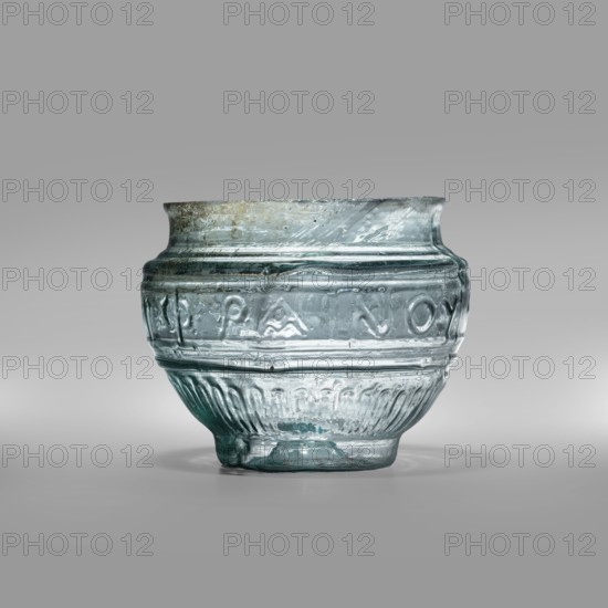 Bowl with Greek inscription. Glass bowl of transparent light blue glass with uneven rim edge, cold-worked. Relief decoration in the lower register: radially arranged long leaves flutes. In the upper, widest part of the bowl there is a Greek inscription: Rejoice that you have come, bounded at the top and bottom by horizontal ribs. Blown in a three-part form. Type: Harden group G.1.IIa. Date: 1st century. Place of origin: Sidon; place of discovery: Cyprus. Vessel; weight 0.070 kg; diameter 4.5 cm bottom, diameter 7.4 cm rim; height 7 cm., A translucent glass vessel with a rounded body and a slightly flared rim sits against a neutral gray background, showing colors of pale blue, aqua, and hints of green with subtle clear areas; the glass surface has a textured appearance with horizontal ridges and a band of raised decorative motifs around the midsection, while the lower portion features vertical fluting and an irregular, slightly uneven foot; small bubbles and surface imperfections are vi...