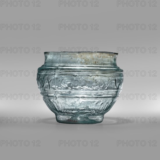 Bowl with Greek inscription. Glass bowl made of transparent, light blue glass with a Greek inscription reading Rejoice that you have come. Uneven rim edge, cold-worked. Relief decoration in the lower register: radially arranged long leaves flutes; inscription in the upper, widest part, bounded at top and bottom by horizontal ribs. Produced by blowing technique in a three-part form. Type: Harden group G.1.IIa. Date: 1st century Sidon; 1st half of the 19th century note appears conflicting. Place of origin: Sidon; place of discovery: Cyprus. Vessel; weight 0.070 kg; diameter 4.5 cm bottom, diameter 7.4 cm rim; height 7 cm., A single translucent glass vessel with a rounded body and slightly flared rim sits against a neutral gray background, showing rippled and irregular surface textures and molded horizontal bands and decorative raised patterns around its midsection; the glass displays a pale blue-green tint with areas of cloudy white and subtle brownish discoloration near the rim, and a s...
