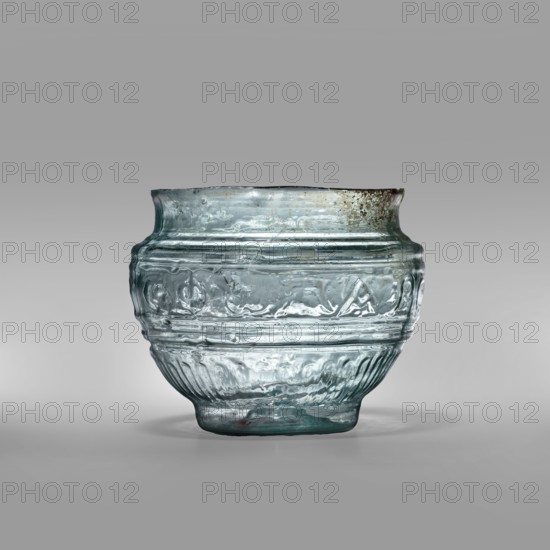 Bowl with Greek inscription. Glass bowl made of transparent, light blue glass with a Greek inscription reading Rejoice that you have come. Uneven rim edge, cold-worked. Relief decoration in the lower register: radially arranged long leaves flutes; inscription in the upper, widest part of the bowl bounded at top and bottom by horizontal ribs. Blown in a three-part form. Type: Harden group G.1.IIa. Date: 1st century or 1st half of the 1st century?. Probable origin: Sidon; findspot: Cyprus. Vessel; weight 0.070 kg; diameter 4.5 cm bottom, diameter 7.4 cm rim; height 7 cm., A translucent glass bowl with a rounded body and a slightly flared rim sits centered on a smooth surface; the glass shows a pale bluish-green tint with areas of clear translucency and some darker green and grayish discoloration near the rim, and the surface of the bowl features embossed horizontal bands and textured decorative patterns including rippled and raised motifs, with a few small bubbles and irregularities in t...