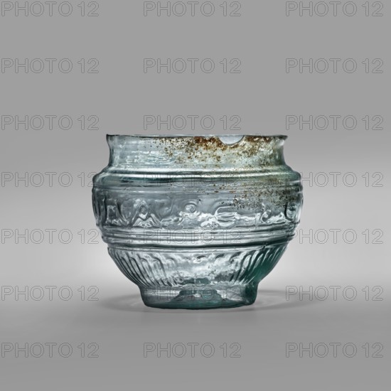 Bowl with Greek inscription. Glass bowl of transparent light blue glass with an uneven rim edge, cold-worked. Relief decoration in the lower register: radially arranged long leaves flutes. In the upper, widest part of the bowl there is a Greek inscription: Rejoice that you have come, bounded at the top and bottom by horizontal ribs. Blown in a three-part form. Type: Harden group G.1.IIa. Date: 1st century manufacture in Sidon; 1st half of the 19th century noted classification. Origin: Sidon; Cyprus discovery. Vessel, weight 0.070 kg; diameter 4.5 cm bottom, diameter 7.4 cm rim, height 7 cm., A translucent glass bowl with a rounded body and a slightly flared rim sits against a neutral gray background; the glass shows a few areas of greenish-blue tint and several patches of brownish discoloration near the rim, with visible air bubbles and irregularities in the surface; the body features a band of embossed decorative motifs and a lower section with vertical ribbing, and the glass rests on...