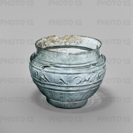 Glass bowl with Greek inscription. Transparent light-blue glass, uneven rim edge, cold-worked. Relief decoration in the lower register: radially arranged long leaves flutes. In the upper, widest part of the bowl a Greek inscription reads Rejoice that you have come, bounded at top and bottom by horizontal ribs. Blown in a three-part form. Type: Harden group G.1.IIa. Dated to 1st century 1st half of the 1st century?. Origin: Sidon; found in Cyprus. Vessel. Weight 0.070 kg; diameter 4.5 cm bottom, diameter 7.4 cm rim; height 7 cm., A translucent glass bowl with a rounded profile sits on a neutral background; the glass shows a pale blue-green tint and areas of cloudiness and surface iridescence, with a few brownish deposits or speckles along the rim and interior edge. The bowl's rim is uneven with a small notch and the glass surface displays several horizontal molded ridges and decorative raised bands encircling the body. The lower portion features a pattern of vertical fluted impressions ...