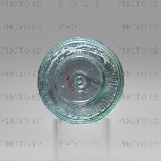 Bowl with Greek inscription. Glass bowl made of transparent, light blue glass with a Greek inscription reading Rejoice that you have come. Uneven rim edge, cold-worked. Relief decoration in the lower register: radially arranged long leaves flutes; inscription in the upper, widest part of the bowl bounded at top and bottom by horizontal ribs. Blown in a three-part form. Harden group G.1.IIa. Dated 1st century Sidon, 1st half of the 19th century note: conflicting dates in original. Origin: Cyprus. Vessel. Weight 0.070 kg; diameter 4.5 cm bottom, diameter 7.4 cm rim; height 7 cm., A round translucent glass object with a bluish-green tint sits centered on a clear cylindrical stand against a neutral gray background; the glass shows concentric circular ridges and a slightly raised central bump, with textured irregularities and small bubbles visible within the material, and a small area of red color near the inner circle.