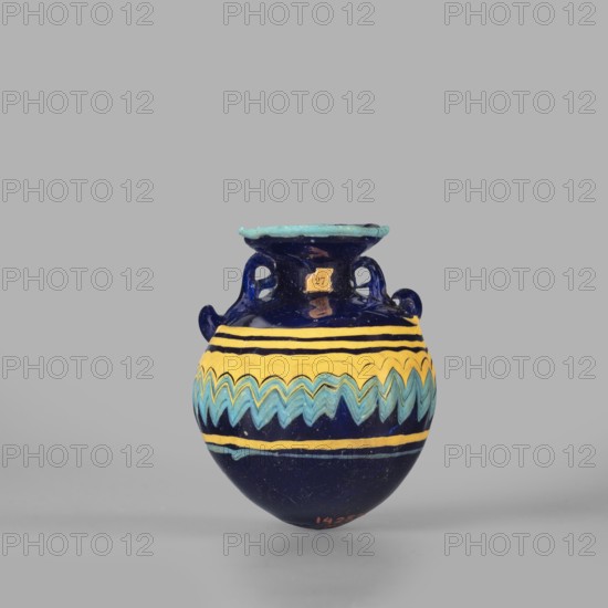 Globular aryballos, Egyptian workshop attributed, globular aryballos decorated at the widest point of the body with a vertically grooved band of zigzag decoration changing into spirals at the bottom and top of the body. Navy blue glass; thread decoration, embedded blue and yellow. 6th–4th century BC., Aryballos vessel; weight 0.05 kg; height 6.5 cm; diameter 5.6 cm., A small rounded ceramic vessel with a flared rim and a pair of small looped handles sits against a neutral gray background, showing a glossy deep blue body with bands of bright yellow and teal decorative zigzag and stripe patterns around its widest part; the rim has a lighter turquoise edge and the handles and upper neck are the same deep blue, with visible glaze reflections and subtle surface wear.