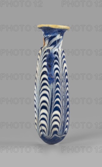Alabastron, Egyptian workshop, 6th–5th century BC. Opaque dark blue glass alabastron decorated with yellow-brown zigzag threads; ovoid body with rounded bottom, flat discoidal rim, and vertical duck vices glued to the upper part of the body; formed on the core. Type: Grose group I:E, form I:6, Harden group I form 4. Height 8.6 cm; body diameter 2.5 cm; spout diameter 2.45 cm; weight 0.030 kg. Site of origin: Egypt, A tall, narrow vase with a flared rim, decorated in alternating curved stripes that create a scalloped, wave-like pattern; the surface shows a glossy finish with raised drip-like accents on one side. The visible colors are deep blue, creamy white, pale yellow on the rim, and small areas of tan. The background is a uniform gray.