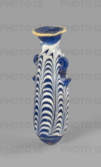 Alabastron, Egyptian workshop, 6th–5th century BC. Opaque dark blue glass alabastron decorated with yellow-brown zigzag threads; ovoid body, rounded bottom, flat discoidal rim, vertical duck handles applied to the upper part of the body; formed on the core. Type: Grose group I:E, form I:6; Harden group I form 4. Dimensions: height 8.6 cm, body diameter 2.5 cm, spout diameter 2.45 cm, weight 0.030 kg. Origin: Egypt, A tall narrow vase displayed against a plain gray background with a flared rim edged in a yellowish-beige color, the body showing a repeating feathered wave pattern in deep blue and white, with molded looped handles on the sides in blue, and the base area continuing the blue and white pattern.