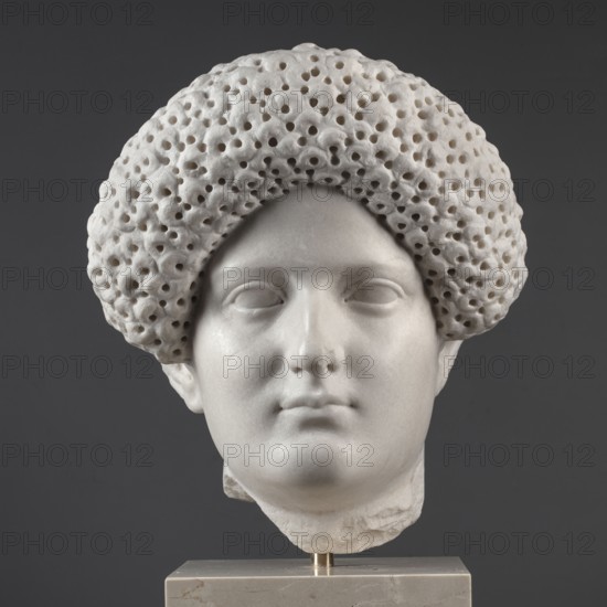 Portrait of a Roman woman, Head with a broad face and an elaborate hairstyle: hair twisted into small curls marked with a gimlet; the rest gathered at the back in a small bun. Round, full face with a triangular forehead. Roman sculpture, female portrait, Roman Empire 27 BC–476, dated 82–92?; sculpture. Dimensions: height 30.5 cm, width 27 cm, depth 25 cm; weight 36.5 kg., A light-colored marble sculpted head displayed against a dark gray background, showing a face with smooth features, closed or shallowly carved eyes, a straight nose, and small lips, topped by a textured rounded hairpiece with a porous, hole-filled surface; the sculpture rests on a pale stone pedestal with a small metallic support visible.