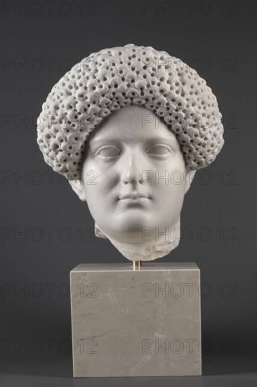 Portrait of a Roman woman, Head with a broad face and an elaborate hairstyle: hair twisted into small curls marked with a gimlet; the rest gathered at the back in a small bun. Round, full face, with a triangular forehead. Roman sculpture, female portrait, masterpiece, hairstyle, dated 82–92, Rome, Roman Empire 27 BC–476. Sculpture. Height 30.5 cm, width 27 cm, depth 25 cm, weight 36.5 kg., A marble sculpture head mounted on a square stone base is centered against a dark gray background; the head is white with smooth facial features and a rounded textured hairpiece that has a porous, hole-filled appearance; the base is a light beige-gray with subtle veining; visible colors include white, light beige-gray, dark gray, and hints of warm beige in the base.