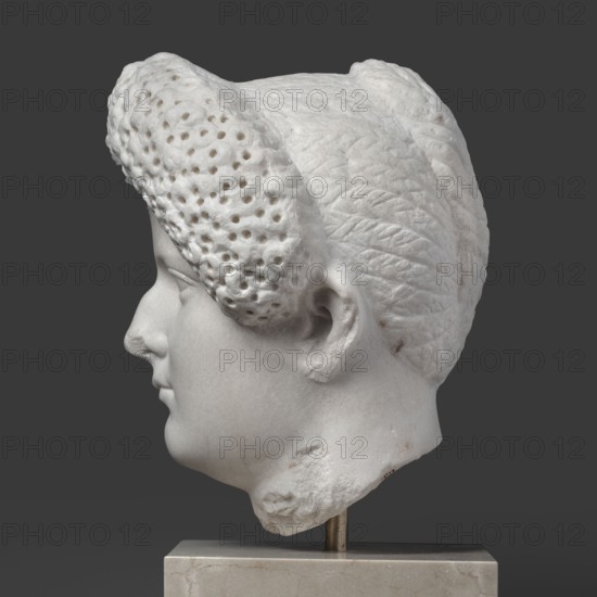 Portrait of a Roman woman, female portrait, Roman sculpture, hairstyle: head with a broad, round full face and a triangular forehead; hair twisted into small curls marked with a gimlet, the rest gathered at the back in a small bun. Masterpiece, dated 82–92, Rome, Roman Empire 27 BC–476. Sculpture. Height 30.5 cm, width 27 cm, depth 25 cm, weight 36.5 kg., A white marble sculpted head in profile set against a dark gray background, showing smooth facial features and a carved ear, with textured hair that includes a patterned, perforated frontal section and braided details at the back; the head rests on a pale beige rectangular base with a metal support, and there are small brownish discolorations on parts of the marble.